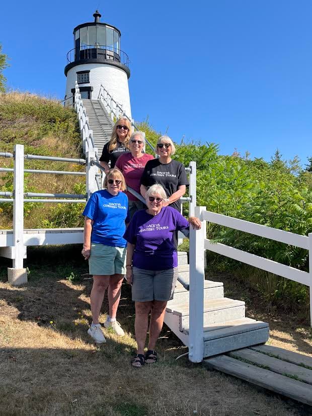 Four women pose together wearing there Dacey's Cornish tours t-shirts on the steps of a lighthouse, smiling and enjoying their time outdoors.