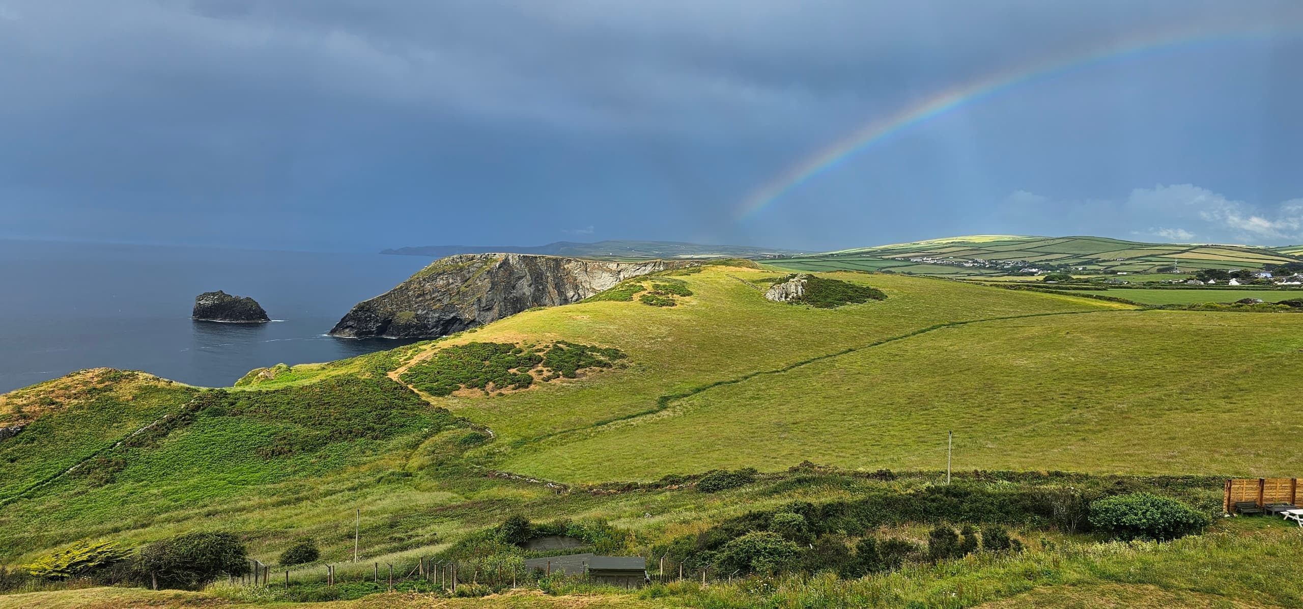 A rainbow over green clifftop fields and a rocky headland under a stormy sky