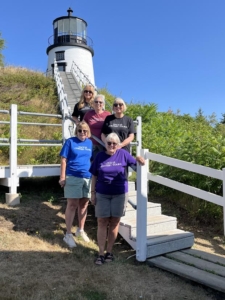 Four women pose together wearing there Dacey's Cornish tours t-shirts on the steps of a lighthouse, smiling and enjoying their time outdoors.